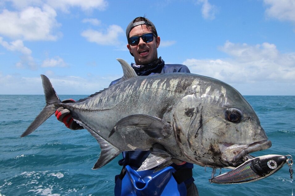 Giant Trevally fish held by a male angler wearing a cap and sunglasses